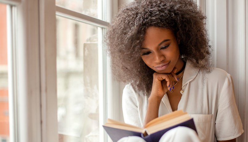 Mulher negra lendo livro sentada ao lado de uma janela