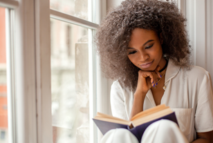 Mulher negra lendo livro sentada ao lado de uma janela